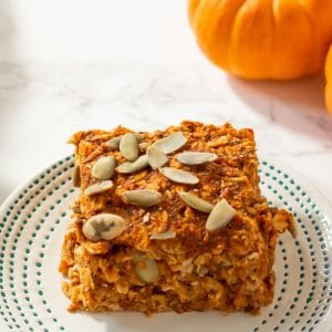 A square of baked pumpkin oatmeal topped with pepitas on a small ivory plate with tiny teal dots on the border.