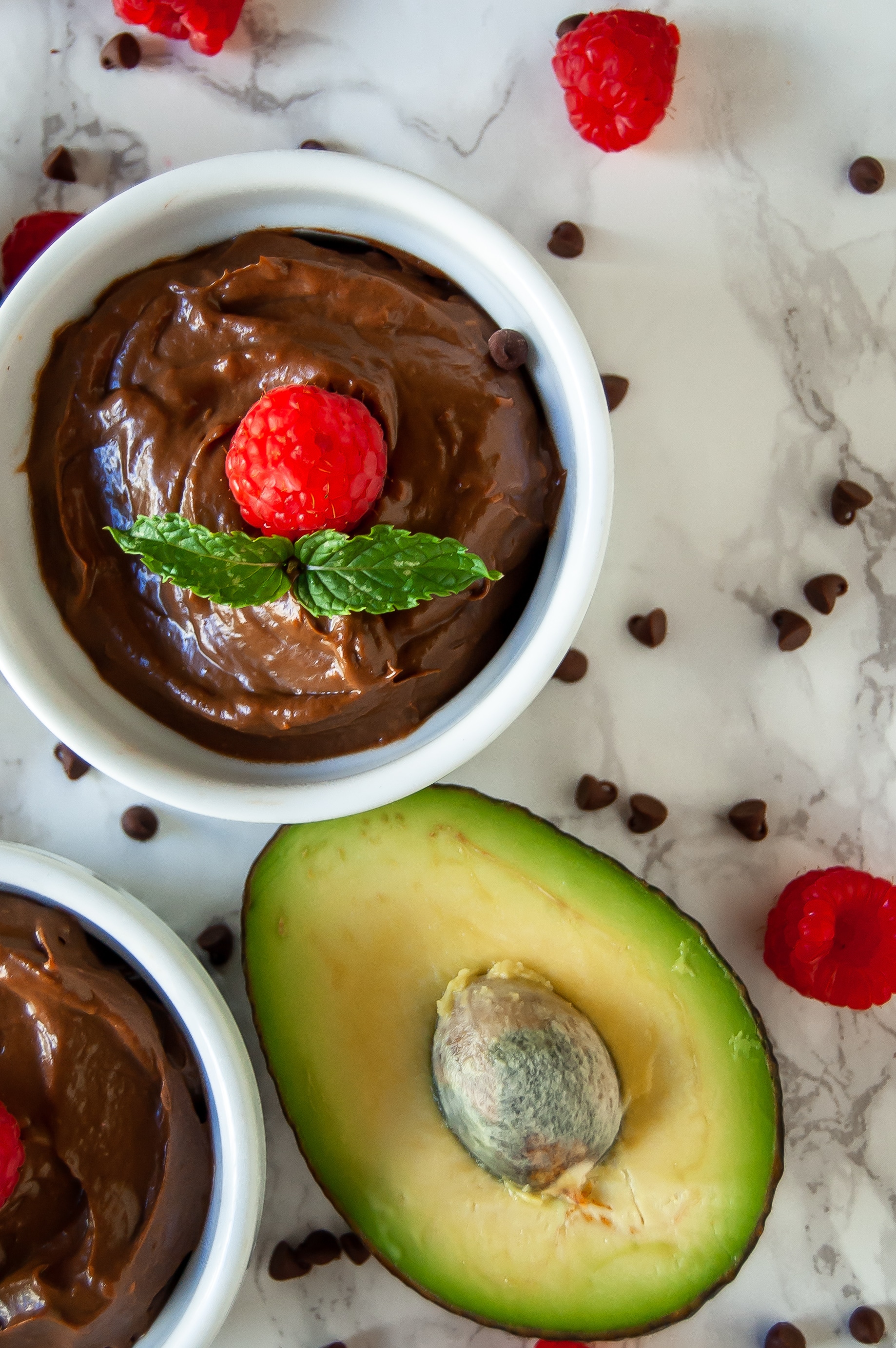Two white ramekins of chocolate raspberry avocado mousse surrounded by avocados, fresh raspberries, and mini chocolate chips.