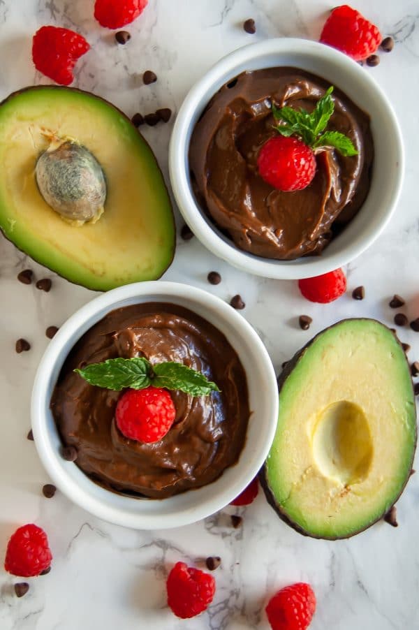 Two white ramekins of chocolate raspberry avocado mousse surrounded by avocados, fresh raspberries, and mini chocolate chips.