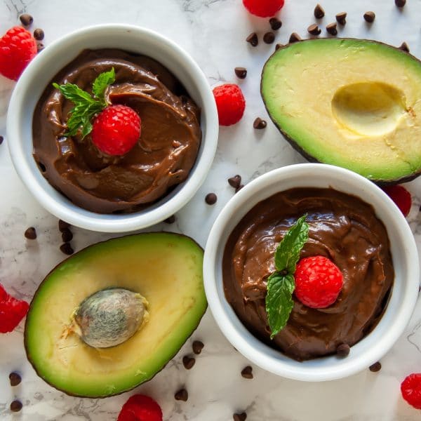 Two white ramekins of chocolate raspberry avocado mousse surrounded by avocados, fresh raspberries, and mini chocolate chips.