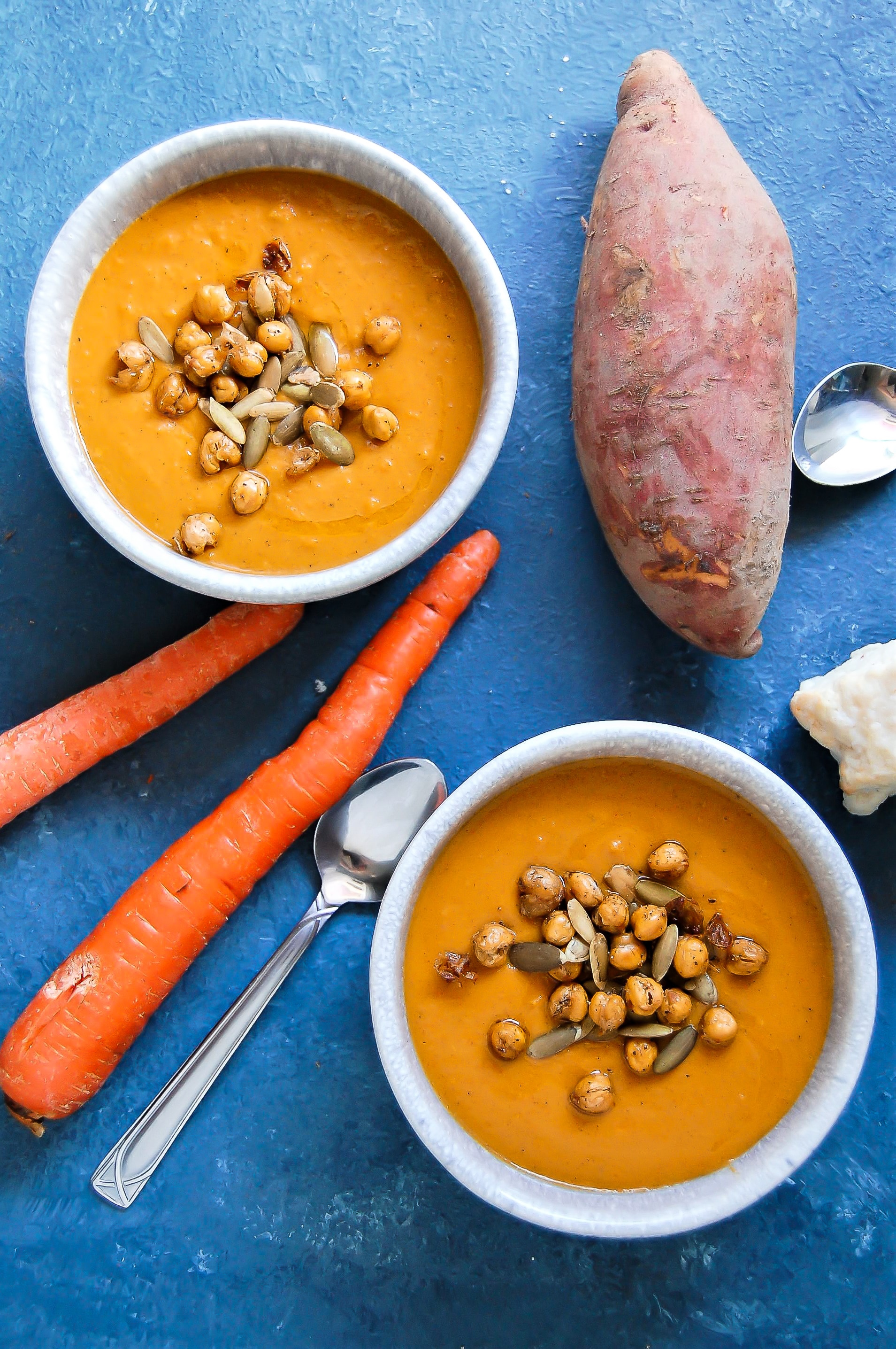 Overhead shot of two bowls of golden orange spiced sweet potato carrot soup garnished with crunchy chickpeas and pumpkin seeds with a sweet potato and carrots on the side on a blue background.