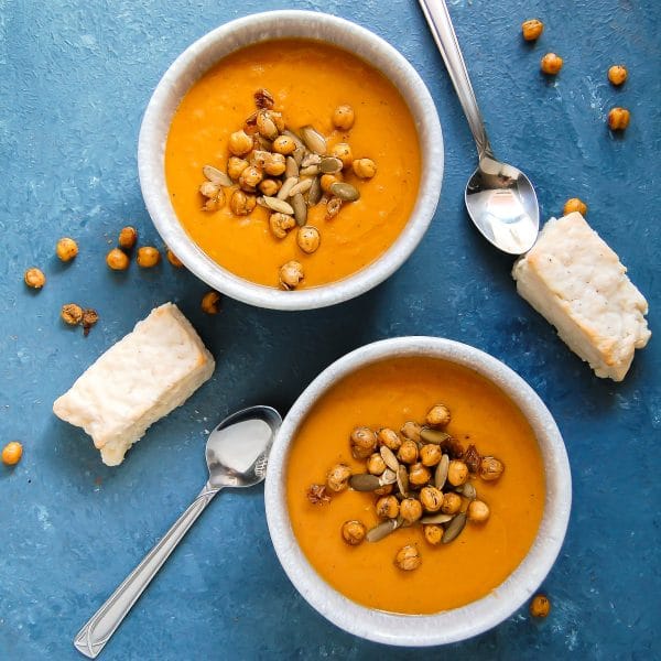 Square image of two bowls of spiced sweet potato carrot soup on a blue background with rectangular biscuits.