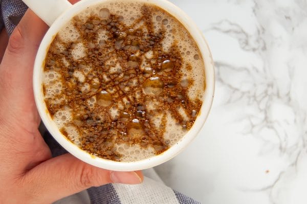 A warm ivory mug with a cozy gingerbread chai latte with molasses drizzle on top being held in a left hand.