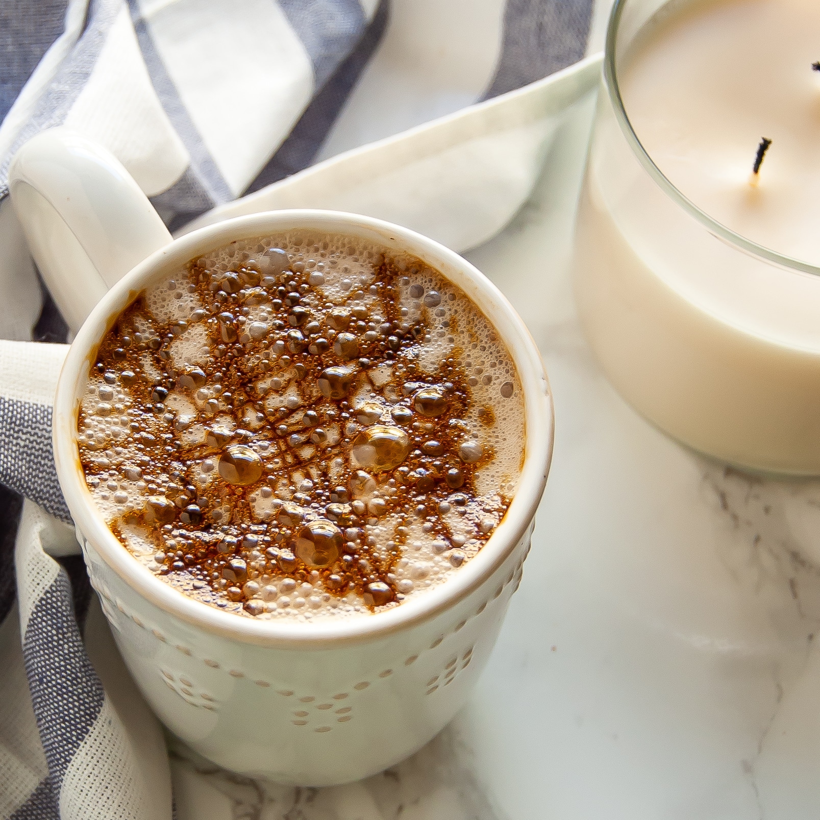 A warm ivory mug with a cozy gingerbread chai latte with molasses drizzle on top next to a candle.