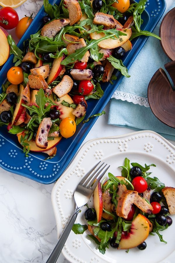 Blueberry Panzanella Salad with Canyon Bakehouse Gluten Free Blueberry Bagels on a blue serving platter and a portion on a white plate.