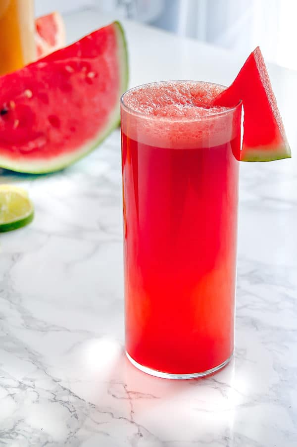 Tall glass of watermelon agua fresca with watermelon and limes in the background.