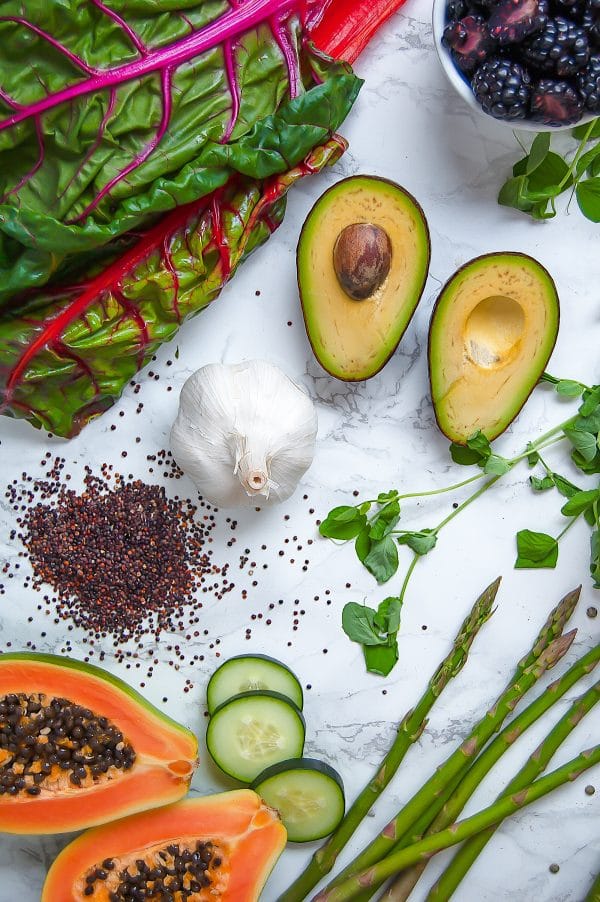 Various plant-based foods, like avocado, garlic, quinoa, pea shoots, asparagus, cucumber, swiss chard, and papaya, displayed beautifully on marble.