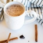 A white mug of chocolate chai tea with cinnamon sticks and dark chocolate on a white board.