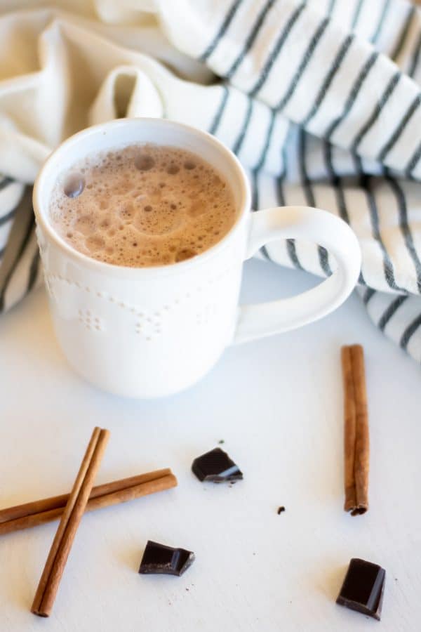 A white mug of chocolate chai tea with cinnamon sticks and dark chocolate on a white board.