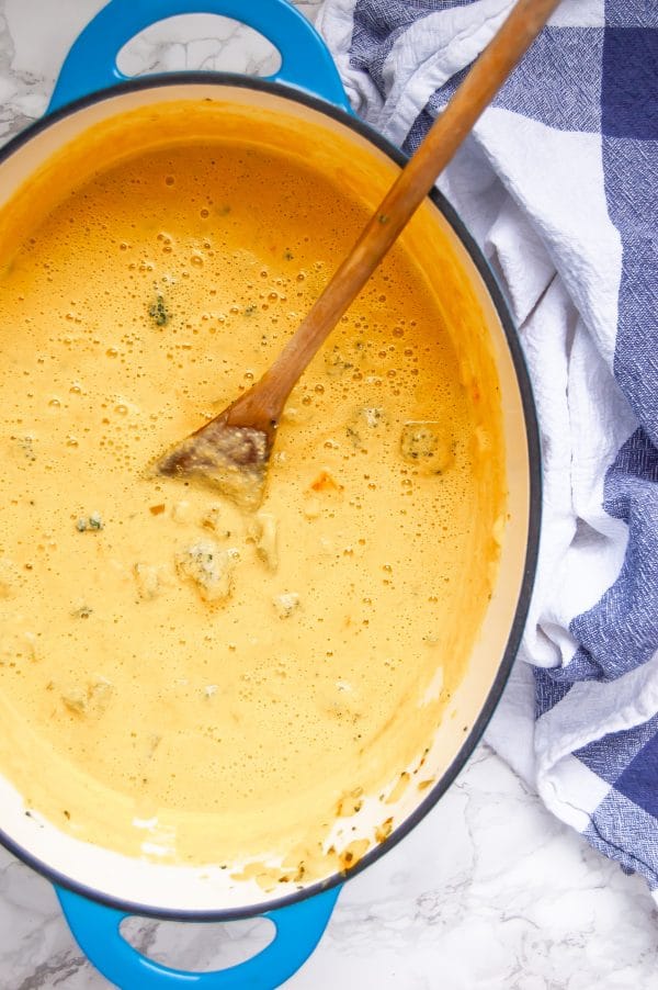 Oval, blue dutch oven full of creamy yellow vegan broccoli cheese soup with a wooden spoon.
