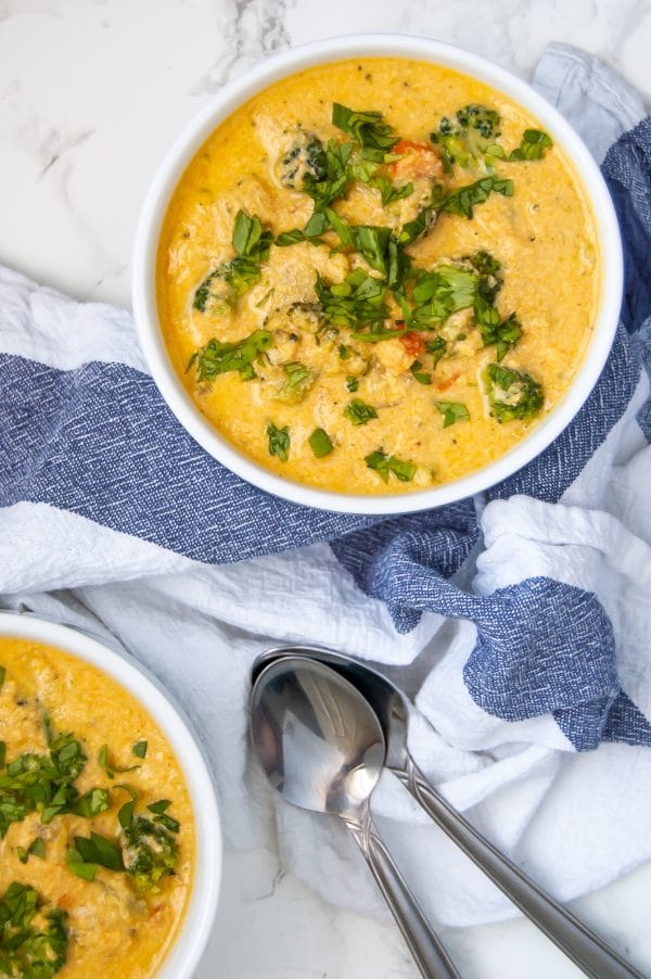 Overhead Shot of white bowls of creamy, golden vegan broccoli cheese soup with herb garnish, surrounded by a white and blue kitchen towel and soup spoons.