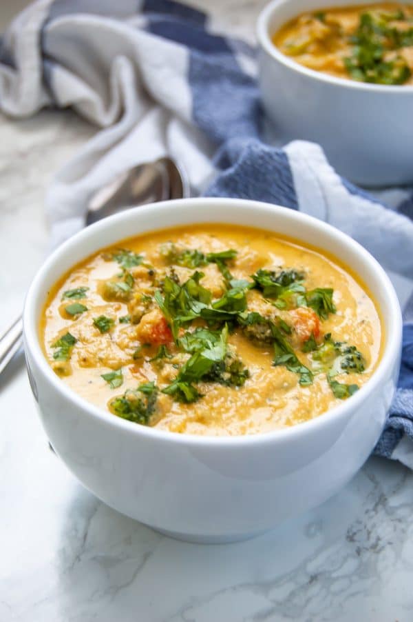 A 45-degree angle shot of golden creamy vegan broccoli cheese soup in a white bowl with an herb garnish. A white and blue towel is in the background