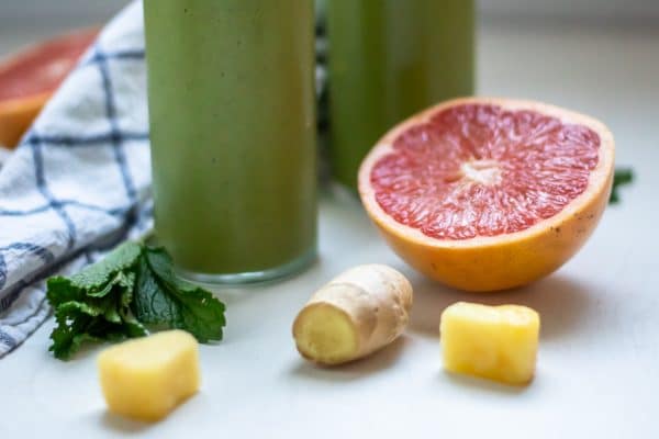 A horizontal image showing the bottom of two glasses of grapefruit smoothies, focused on the fresh grapefruit, ginger, and pineapple ingredients on the sides.