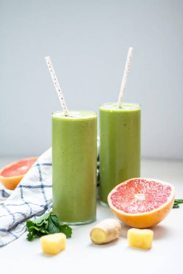 Two tall, thin glasses of green grapefruit smoothies with paper straws, flanked by fresh grapefruit, pineapple, ginger, and mint.