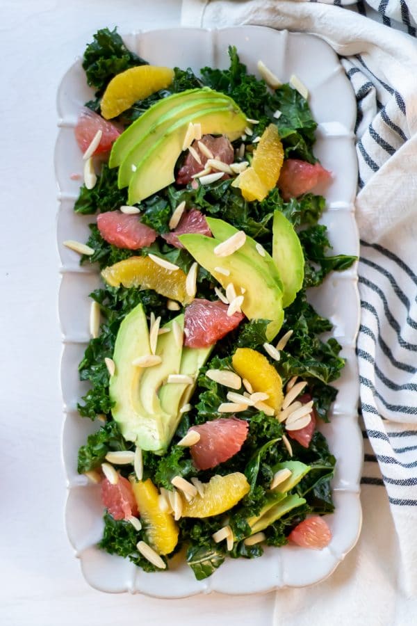 Rectangular plate full of a citrus kale salad massaged with grapefruit vinaigrette and topped with sliced avocado, citrus segments, and slivered almonds.