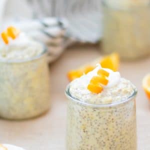 3 small glasses of orange chia pudding with collagen on a burlap table runner with orange slices. Garnished with coconut whipped cream and curled orange peel garnishes.