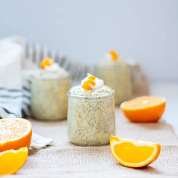 3 small glasses of orange chia pudding with collagen on a burlap table runner with orange slices. Garnished with coconut whipped cream and curled orange peel garnishes.