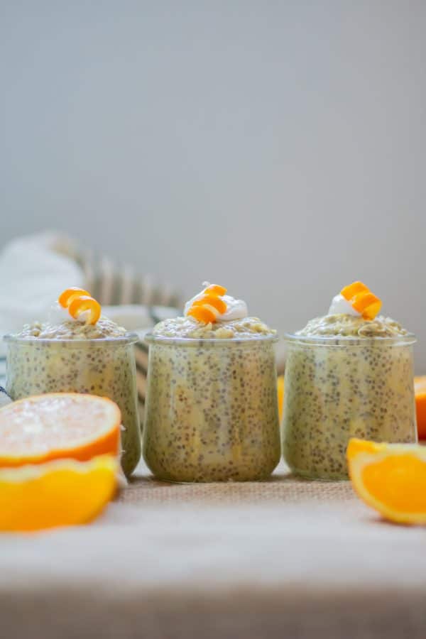 3 small glass jars of orange chia pudding with collagen lined up next to each other topped with coconut whipped cream and orange curls.