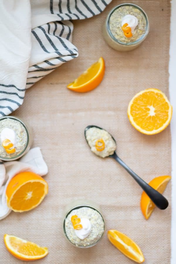 Glass cups of orange chia pudding with a black spoon with a spoonful of chia pudding, accented by orange slices all on a burlap runner.