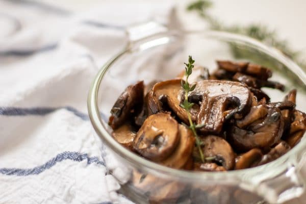 Close up of healthy, easy sauteed mushrooms in a glass bowl garnished with a sprig of thyme.