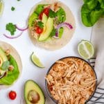 A glass bowl of instant pot salsa chicken flanked by tacos and taco toppings.