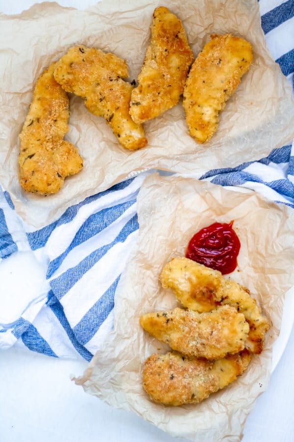 Overhead shot of a platter of garlic parmesan chicken tenders and a parchment covered plate with more tenders and ketchup on a white board with a blue striped linen towel.