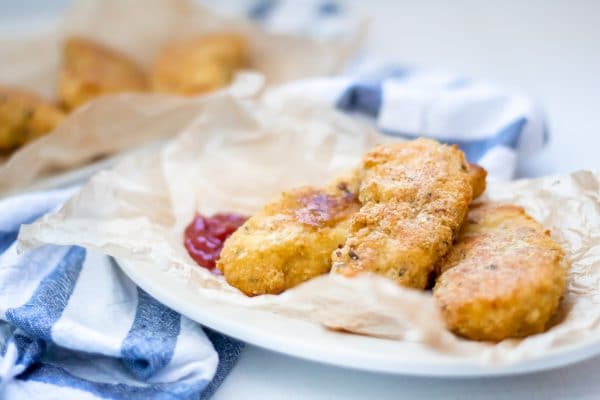 A white plate covered with brown parchment paper holding garlic parmesan chicken tenders and ketchup.