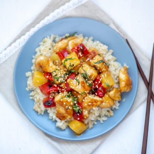 A blue plate full of cauliflower rice topped with healthy sweet and sour chicken and garnished with fresh herbs.