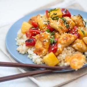 A blue plate full of cauliflower rice topped with healthy sweet and sour chicken and garnished with fresh herbs. It's sitting on a light brown linen napkin with brown chopsticks by the plate.