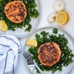 Two salmon patties on plates of massaged kale salad with garlic and lemon on the side.
