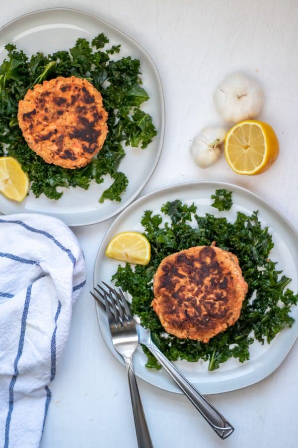 Two salmon patties on plates of massaged kale salad with garlic and lemon on the side.