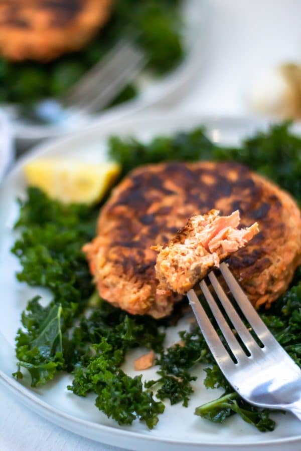 A forkful of salmon patty from a salmon patty on a bed of massaged kale with a lemon wedge on the side.