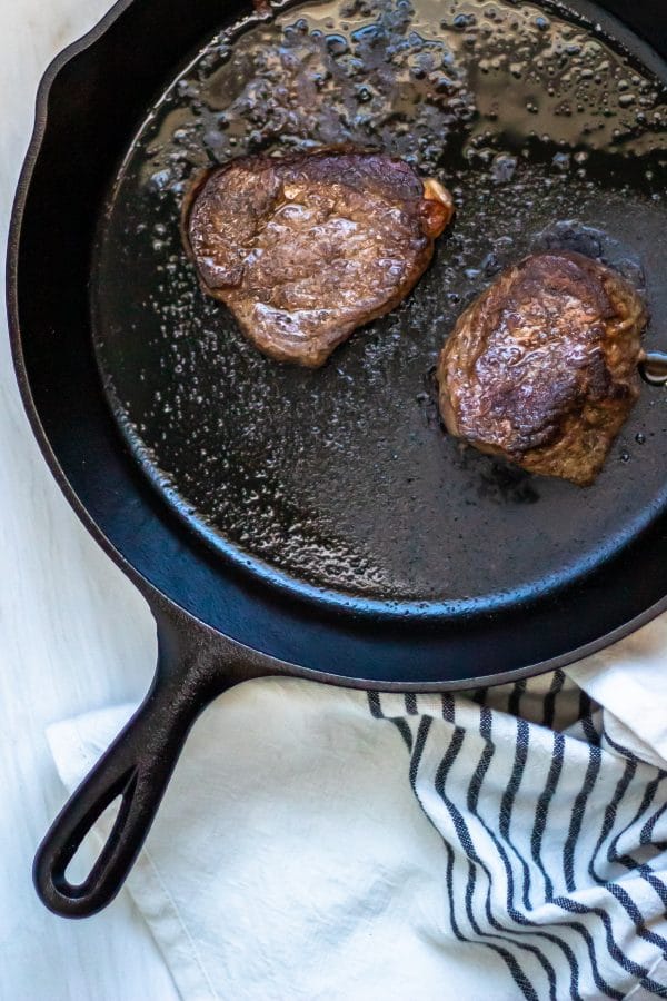 Two seared sirloin steaks in a cast iron skillet next to a cream and black striped linen kitchen towel.