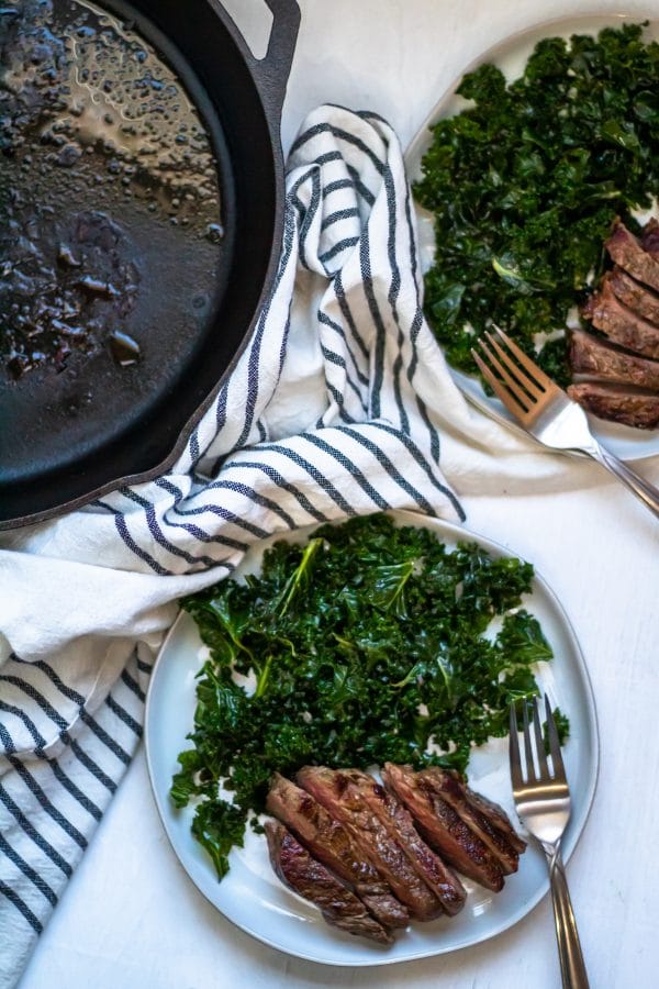 Two white plates with sliced steak and massaged kale salad next to a cast iron skillet.