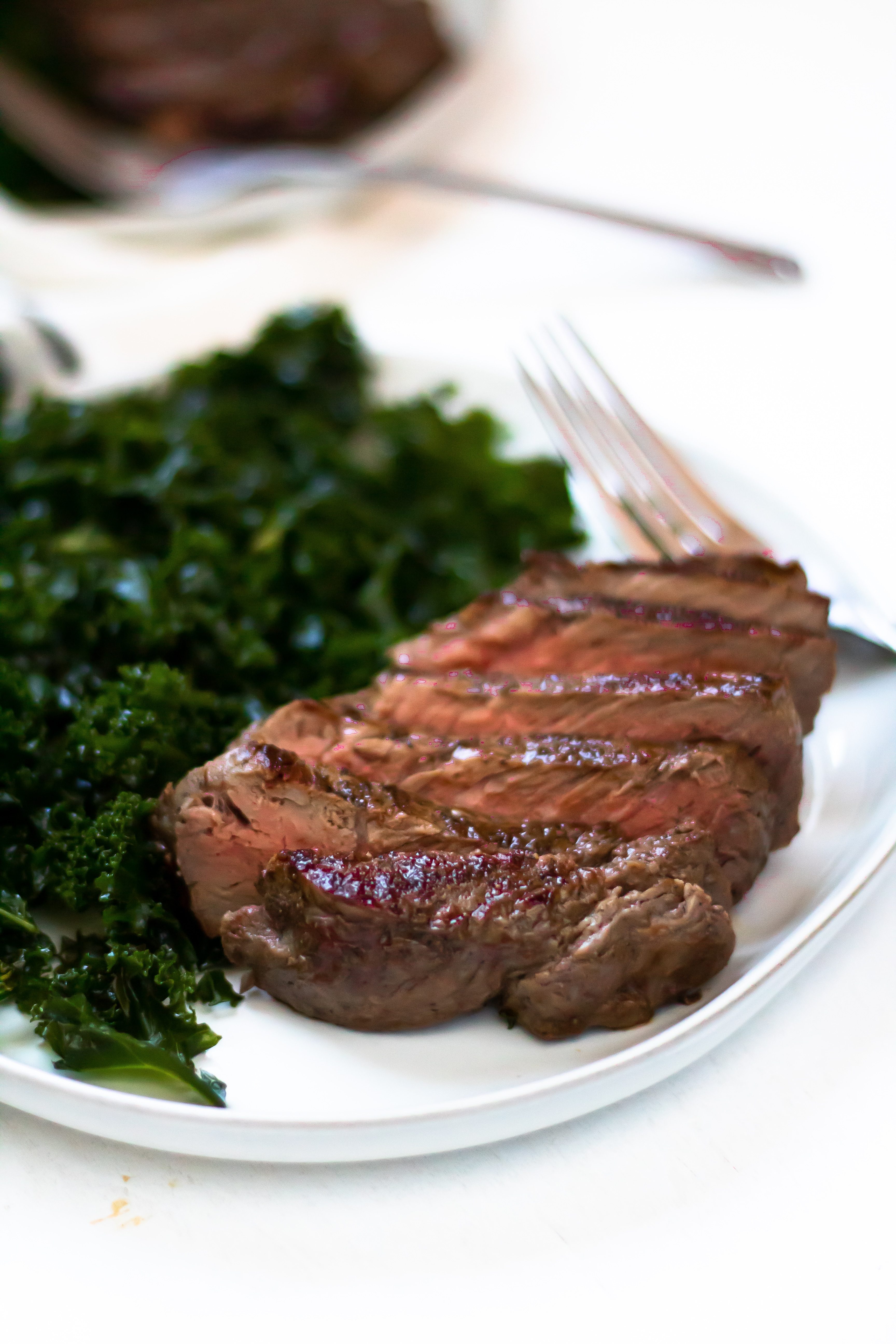 A seared and sliced cast iron skillet steak on a white plate with massaged kale salad.