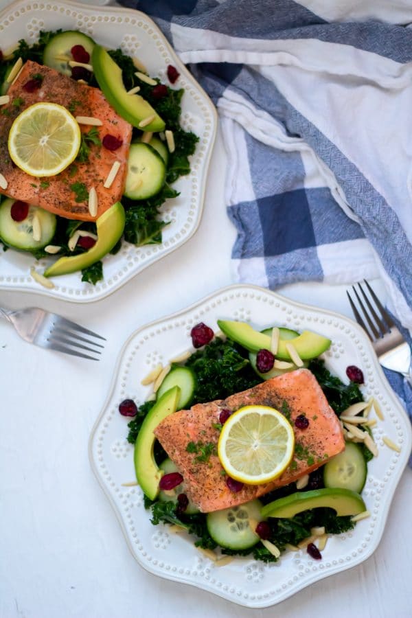 Overhead shot of two kale salads topped with salmon and lemon wedges with a white and blue dish towel to the side on a white wooden board.