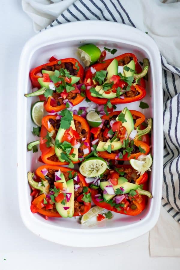 Red bell peppers stuffed with turkey filling for taco stuffed peppers in a white casserole dish, garnished with fresh toppings like avocado, lime, tomato, red onion, and cilantro.