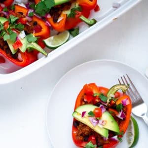 A taco stuffed red bell pepper on a white plate with white background topped with fresh avocado, tomato, cilantro, lime, and red onion.