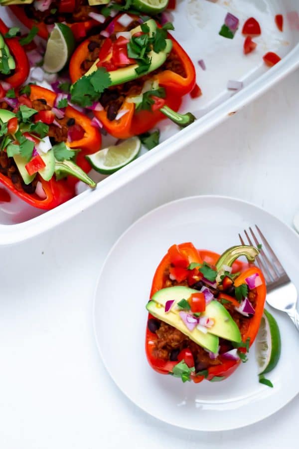 A taco stuffed red bell pepper on a white plate with white background topped with fresh avocado, tomato, cilantro, lime, and red onion.