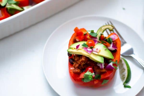 A taco stuffed red bell pepper on a white plate with white background topped with fresh avocado, tomato, cilantro, lime, and red onion.