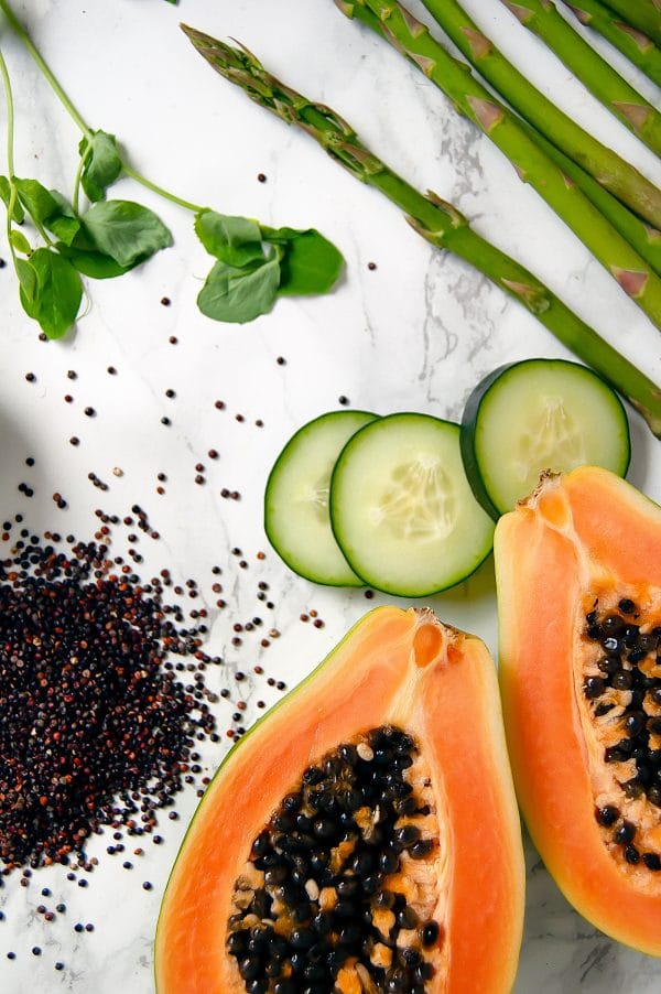 Marble background with an open papaya, cucumber slices, asparagus, dried red quinoa, and pea tendrils.
