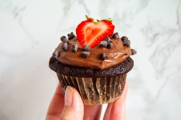 A hand holding a dark chocolate cupcake with chocolate frosting topped with mini chocolate chips and half a strawberry.