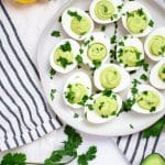 Cream ceramic plate with avocado deviled eggs garnished with cilantro on a black and cream striped linen towel and lemons, avocado, and cilantro surrounding it.