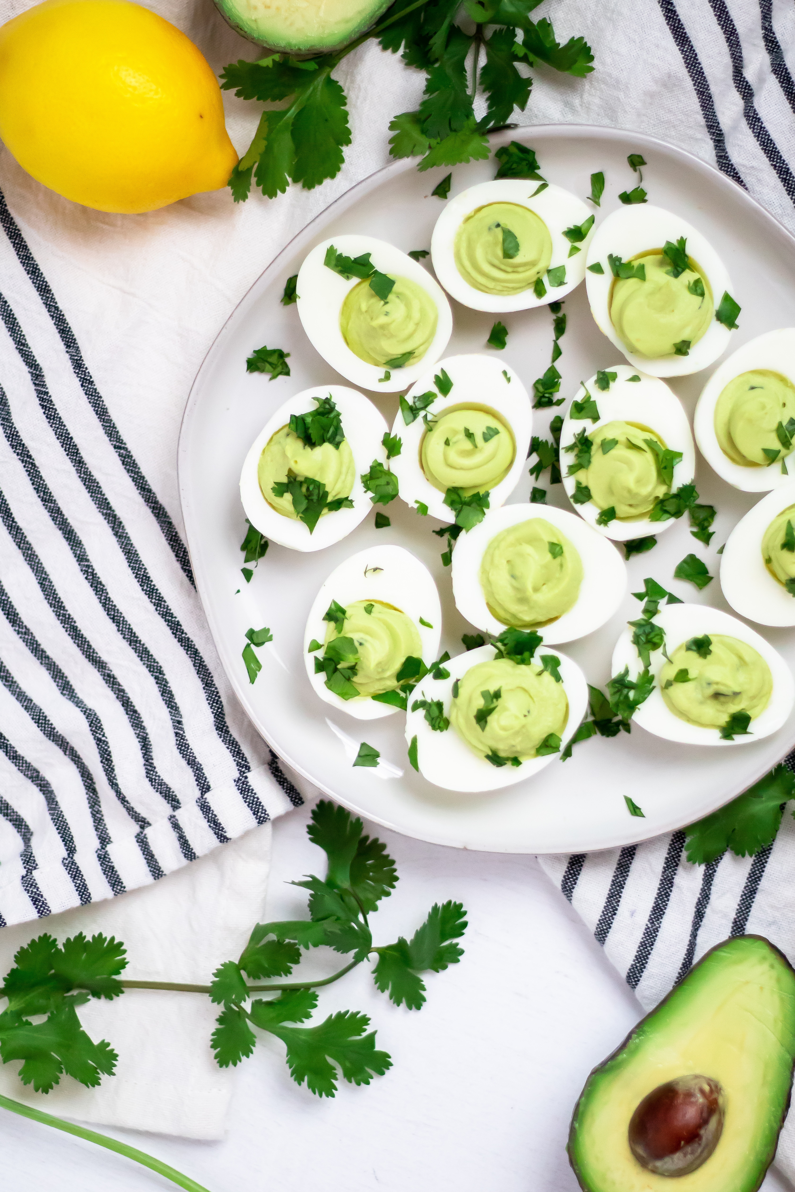 Cream ceramic plate with avocado deviled eggs garnished with cilantro on a black and cream striped linen towel and lemons, avocado, and cilantro surrounding it.