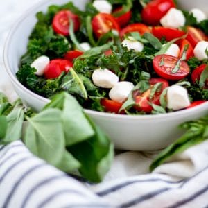 Kale caprese salad in a cream ceramic shallow owl surrounded by fresh basil and tomatoes.