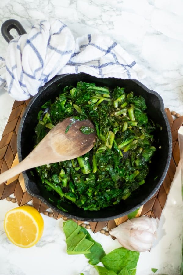 Cast iron skillet of sauteed lemon garlic turnip greens with a wooden spoon and white and blue towel over the skillet handle.