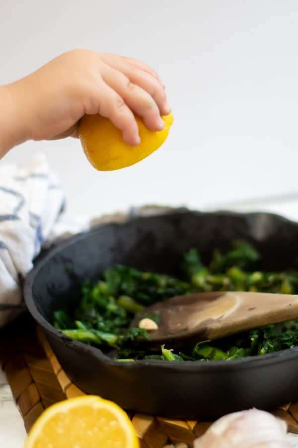A child's hand squeezing half a lemon over a cast iron skillet of turnip greens.