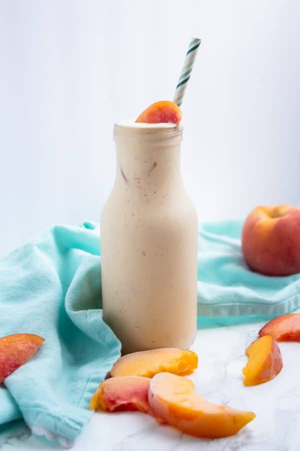 a glass bottle of creamy slightly peachy colored banana peach smoothie with frozen peach slices and a blue cloth behind it.