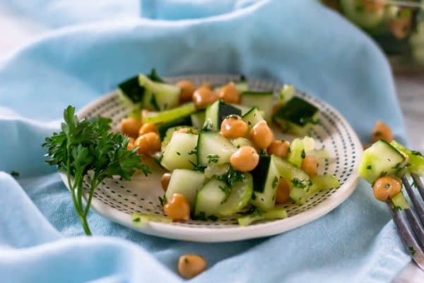 A small plate of cucumber chickpea salad with a sprig of parsley on a light blue cloth.