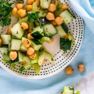 A small white plate with teal dots topped with cucumber chickpea salad with a fork with a bite of salad on it to the side.